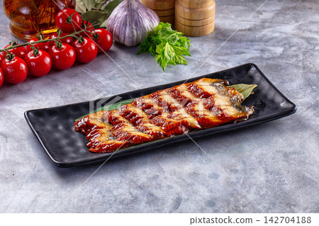Grilled eel with glossy sauce on black rectangular plate, surrounded by cherry tomatoes, garlic, parsley and wooden spice grinders on gray stone surface 142704188