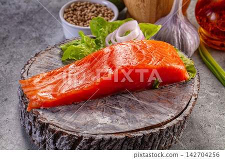 Raw salmon fillet on rustic wooden cutting board with fresh lettuce, sliced onion, garlic, and peppercorns in background Raw salmon fillet on rustic wooden cutting board with fresh lettuce, sliced onion, garlic, and peppercorns in background 142704256