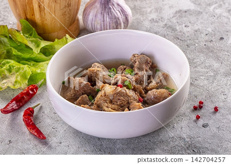 Bowl of tender braised lamb stew with green onions, red chilies, and peppercorns on gray stone surface with garlic and lettuce nearby Bowl of tender braised lamb stew with green onions, red chilies, and peppercorns on gray stone surface with garlic and lettuce nearby 142704257