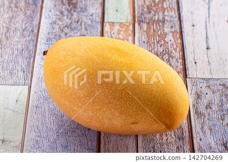 Ripe yellow mango on rustic wooden surface with textured grain and soft natural lighting Ripe yellow mango on rustic wooden surface with textured grain and soft natural lighting 142704269