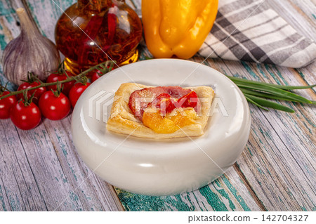 Savory puff pastry tartlet with melted cheese, roasted tomato, and ham on white plate surrounded by fresh garlic, cherry tomatoes, yellow bell pepper, and chives Savory puff pastry tartlet with melted cheese, roasted tomato, and ham on white plate surrounded by fresh garlic, cherry tomatoes, yellow bell pepper, and chives 142704327