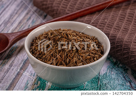 White ceramic bowl filled with brown cumin seeds on rustic wooden surface with wooden spoon and textured cloth background White ceramic bowl filled with brown cumin seeds on rustic wooden surface with wooden spoon and textured cloth background 142704350