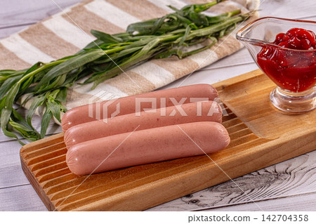 Three raw sausages on wooden cutting board with fresh green herbs and red ketchup in glass bowl on striped cloth background 142704358