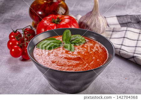 Bowl of vibrant red gazpacho garnished with fresh basil, surrounded by ripe tomatoes, garlic bulb, and olive oil bottle on textured gray surface Bowl of vibrant red gazpacho garnished with fresh basil, surrounded by ripe tomatoes, garlic bulb, and olive oil bottle on textured gray surface 142704368