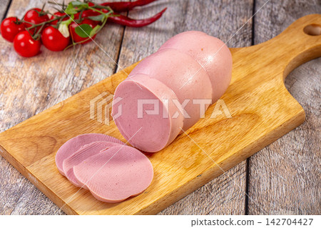 Pink sausage log with slices on wooden cutting board, cherry tomatoes and red chili peppers in background, rustic kitchen setting 142704427