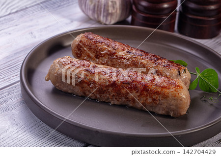 Two golden brown roasted sausages on matte gray plate with fresh green herb garnish, garlic and wooden spice grinders in background 142704429