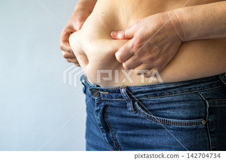 Woman pinching belly fat above denim jeans, body image and weight concern Woman pinching belly fat above denim jeans, body image and weight concern 142704734