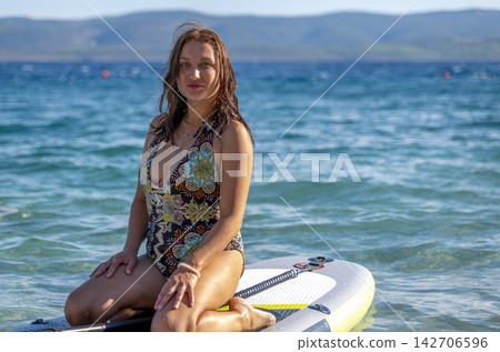 Smiling Woman Sitting on Paddleboard in Calm Water with Mountains Smiling Woman Sitting on Paddleboard in Calm Water with Mountains 142706596