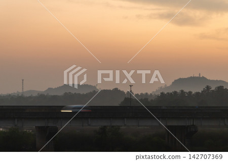 A blurred car crosses an elevated concrete bridge at sunset, while misty silhouetted hills A blurred car crosses an elevated concrete bridge at sunset, while misty silhouetted hills 142707369