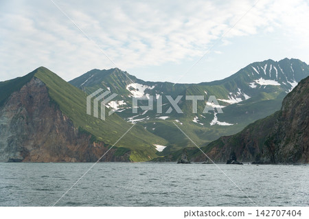 Green mountain ridges and melting snowfields overlook coastal waters in Kamchatka. Remote wilderness, dramatic landscape, scenic marine journey, nature tourism, adventure travel and exploration. 142707404