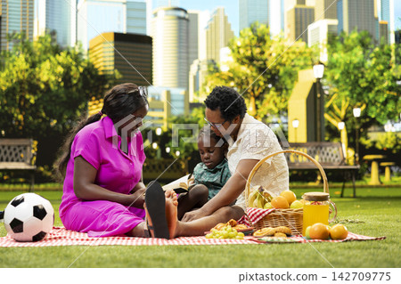African parents and child reading book together on picnic blanket, sharing calm learning and storytelling moment beside food and picnic basket in urban park. Weekend fun. African parents and child reading book together on picnic blanket, sharing calm learning and storytelling moment beside food and picnic basket in urban park. Weekend fun. 142709775