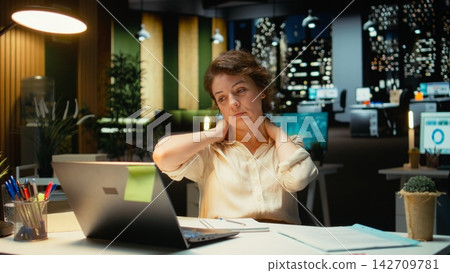 Businesswoman stretching her upper back and neck muscles, correcting the posture and neck stiffness for relief and relaxation. Intense office activity due to overtime late at night. Camera A. Businesswoman stretching her upper back and neck muscles, correcting the posture and neck stiffness for relief and relaxation. Intense office activity due to overtime late at night. Camera A. 142709781