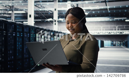 Programmer in data center using laptop to upgrade systems for increased stability. IT professional in server farm configuring infrastructure with notebook for optimum performance, camera A 142709786