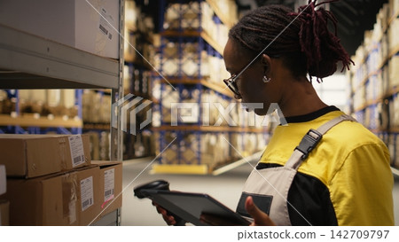 Logistics hub woman employee scanning airway bill tags for registration in inventory, updating records in import export system on tablet. E-commerce operations in industrial depot. Camera A. Logistics hub woman employee scanning airway bill tags for registration in inventory, updating records in import export system on tablet. E-commerce operations in industrial depot. Camera A. 142709797