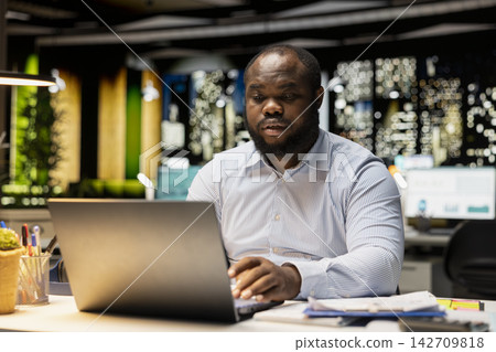 African american guy typing information on laptop and checking reports, does proofreading and reviewing data for a new business objective or target. Company management after hours. African american guy typing information on laptop and checking reports, does proofreading and reviewing data for a new business objective or target. Company management after hours. 142709818