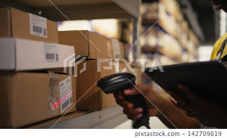 African american woman using scanner to register barcodes tags in inventory, solving logistics operations in industrial warehouse. Worker checking shipping labels near storage racks. Camera A. African american woman using scanner to register barcodes tags in inventory, solving logistics operations in industrial warehouse. Worker checking shipping labels near storage racks. Camera A. 142709819