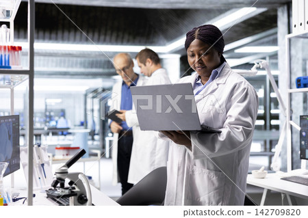 Black female researcher in a clinical research lab studies experiment data with a laptop. Showcasing biotechnology, genetics research and pharmacology for progress in healthcare. Black female researcher in a clinical research lab studies experiment data with a laptop. Showcasing biotechnology, genetics research and pharmacology for progress in healthcare. 142709820