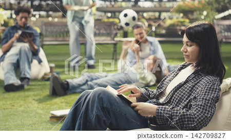 Woman student holding open book seated on beanbag, studying alone outdoors with notebook. Independent learning session on modern campus with soft background blur. Woman student holding open book seated on beanbag, studying alone outdoors with notebook. Independent learning session on modern campus with soft background blur. 142709827