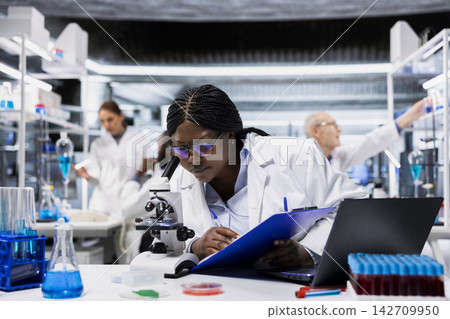 Female microscopist inspects new drug compound under microscope, documenting findings. Lab worker verifying medication quality and purity standards with optical device, writing, taking notes Female microscopist inspects new drug compound under microscope, documenting findings. Lab worker verifying medication quality and purity standards with optical device, writing, taking notes 142709950
