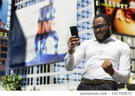 Adult man feeling proud of his new accomplishment on city center street, being excited and cheering on the sidewalk downtown. Pedestrian reading good news on his smartphone. Adult man feeling proud of his new accomplishment on city center street, being excited and cheering on the sidewalk downtown. Pedestrian reading good news on his smartphone. 142709972