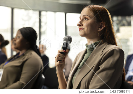 Young woman in attendance asking a question on microphone during conference debate, interacting with speaker and sharing her opinion. International summit with audience engagement. Young woman in attendance asking a question on microphone during conference debate, interacting with speaker and sharing her opinion. International summit with audience engagement. 142709974