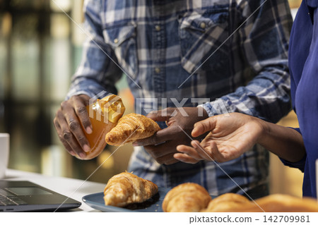 Close up of black couple preparing croissants with honey and fresh toast for delicious breakfast meal in the kitchen. Man and woman prepares homemade food, the joy of a healthy lifestyle. Close up of black couple preparing croissants with honey and fresh toast for delicious breakfast meal in the kitchen. Man and woman prepares homemade food, the joy of a healthy lifestyle. 142709981