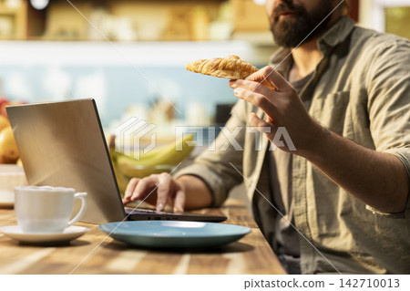 Close up of male eating croissants and working on laptop at breakfast, serving pastry and coffee at the kitchen table. Taking a bite and solving daily tasks, flexible work at home. Close up of male eating croissants and working on laptop at breakfast, serving pastry and coffee at the kitchen table. Taking a bite and solving daily tasks, flexible work at home. 142710013