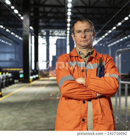 Portrait of worker standing beside heavy machinery in a warehouse facility, wearing safety glasses and a safety vest while overseeing fabrication tools. Manpower in industrial environment. Portrait of worker standing beside heavy machinery in a warehouse facility, wearing safety glasses and a safety vest while overseeing fabrication tools. Manpower in industrial environment. 142710092