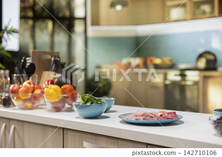 Empty kitchen with wooden dining furniture and modern appliances. Countertop displays colorful vegetables, fresh fruits and spices, representing a stylish home with cozy atmosphere. Empty kitchen with wooden dining furniture and modern appliances. Countertop displays colorful vegetables, fresh fruits and spices, representing a stylish home with cozy atmosphere. 142710106