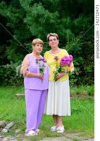 Full length portrait of two senior russian sisters 80 and 75 years old holding flower bouquets standing together in a garden in Russia Full length portrait of two senior russian sisters 80 and 75 years old holding flower bouquets standing together in a garden in Russia 142710473