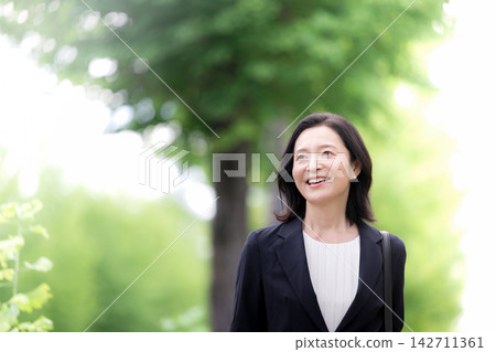 一位五十多歲的女商人漫步在綠樹成蔭的城市中;對於通勤上班的職業婦女來說,這是一種令人耳目一新的生活方式。 一位五十多歲的女商人漫步在綠樹成蔭的城市中;對於通勤上班的職業婦女來說,這是一種令人耳目一新的生活方式。 142711361