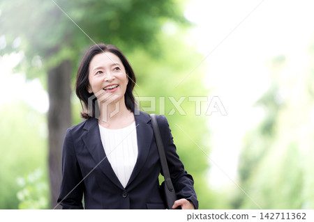 一位五十多歲的女商人漫步在綠樹成蔭的城市中;對於通勤上班的職業婦女來說,這是一種令人耳目一新的生活方式。 一位五十多歲的女商人漫步在綠樹成蔭的城市中;對於通勤上班的職業婦女來說,這是一種令人耳目一新的生活方式。 142711362