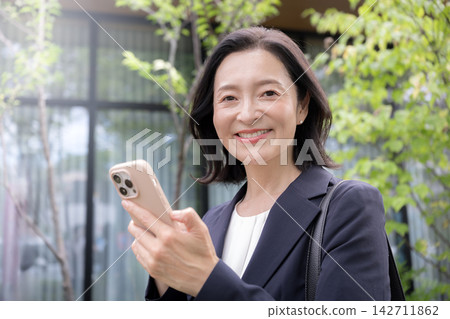 A businesswoman in her 50s operating her smartphone outdoors; a working woman checking messages and map apps while out and about. A businesswoman in her 50s operating her smartphone outdoors; a working woman checking messages and map apps while out and about. 142711862