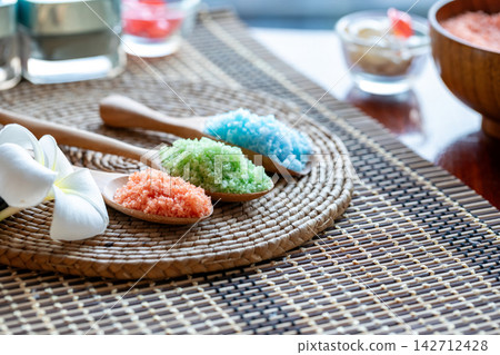 Colorful bath salt crystals in wooden spoons with white frangipani flower on woven mat for spa, wellness, relaxation, beauty, therapy and natural body care 142712428