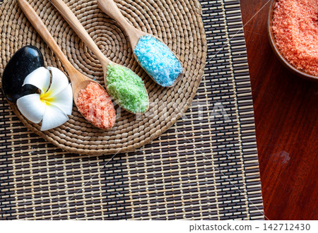 Colorful bath salt crystals in wooden spoons with spa stone, white frangipani flower and woven mat for wellness, relaxation, beauty, therapy and natural body care 142712430