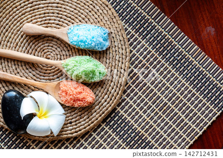Colorful bath salt in wooden spoons with black spa stone and white tropical flower on woven mat for wellness, relaxation, beauty, therapy and natural body care Colorful bath salt in wooden spoons with black spa stone and white tropical flower on woven mat for wellness, relaxation, beauty, therapy and natural body care 142712431