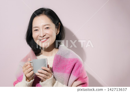 A woman in her 50s, holding a glass of warm beverage against a pink background with both hands, smiles as she enjoys a relaxing winter moment. A woman in her 50s, holding a glass of warm beverage against a pink background with both hands, smiles as she enjoys a relaxing winter moment. 142713637