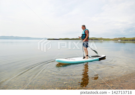 A man in a life jacket rides a paddle board on a lake. Supboard water sport activity A man in a life jacket rides a paddle board on a lake. Supboard water sport activity 142713718