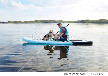 A man sits on a paddle board on a calm lake with a dog beside him. It is sunny and there are green hills in the background. A man sits on a paddle board on a calm lake with a dog beside him. It is sunny and there are green hills in the background. 142713728