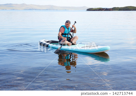 Man paddle boarding on a calm mountain lake wearing a life jacket, enjoying outdoor recreation and water sports  142713744