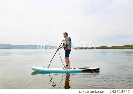 A man in a life jacket rides a paddle board on a lake. Supboard water sport activity A man in a life jacket rides a paddle board on a lake. Supboard water sport activity 142713745