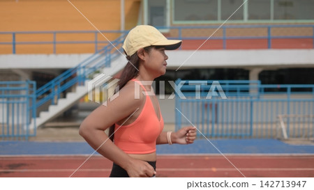 Side view of an active Asian woman wearing a cap and orange sports bra jogging on a running track at an outdoor stadium for fitness training and health lifestyle. Side view of an active Asian woman wearing a cap and orange sports bra jogging on a running track at an outdoor stadium for fitness training and health lifestyle. 142713947