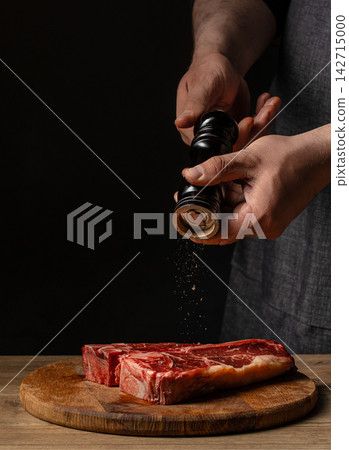 Close-up of hands seasoning raw beef steak with black pepper using a grinder on a wooden cutting board against a dark background Close-up of hands seasoning raw beef steak with black pepper using a grinder on a wooden cutting board against a dark background 142715000