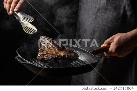 Person holding a cast iron skillet with grilled steak using tongs, smoke rising in the air against a dark background, showcasing culinary preparation and cooking techniques Person holding a cast iron skillet with grilled steak using tongs, smoke rising in the air against a dark background, showcasing culinary preparation and cooking techniques 142715004