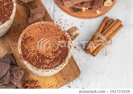 Hot chocolate in a decorative mug topped with cocoa powder, surrounded by chocolate pieces, cinnamon sticks, and star anise on a wooden board Hot chocolate in a decorative mug topped with cocoa powder, surrounded by chocolate pieces, cinnamon sticks, and star anise on a wooden board 142715039