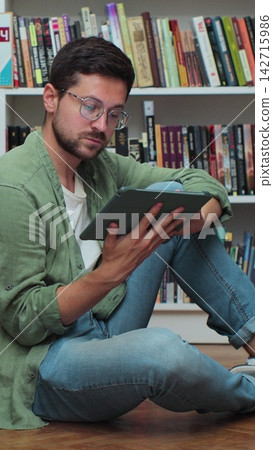 Caucasian man sitting on the floor near shelves with book in the library, uses digital tablet computer and writes notes for class assignment. Caucasian man sitting on the floor near shelves with book in the library, uses digital tablet computer and writes notes for class assignment. 142715986