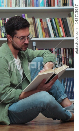 Caucasian student man reading book, sitting among stack of books on library floor, side view. Caucasian student man reading book, sitting among stack of books on library floor, side view. 142715987
