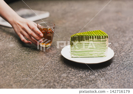Waiter serving a small glass of tea alongside with green tea crepe cake on stone counter. Waiter serving a small glass of tea alongside with green tea crepe cake on stone counter. 142716179