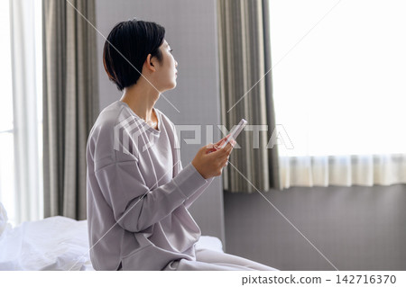 A woman in bed looks at her smartphone and gazes out the window. A woman in bed looks at her smartphone and gazes out the window. 142716370
