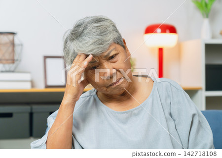 Stressed elderly Asian woman with a headache sits on a sofa at home. Stressed elderly Asian woman with a headache sits on a sofa at home. 142718108